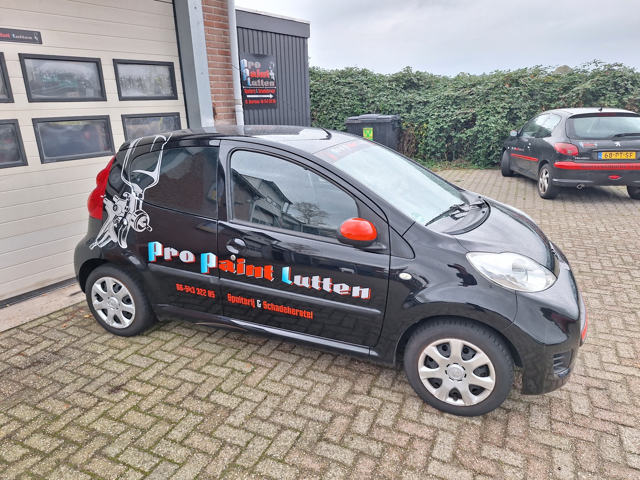 De belettterde bedrijfsauto van Pro Paint Lutten voor de werkplaats, met het bord aan de muur erachter