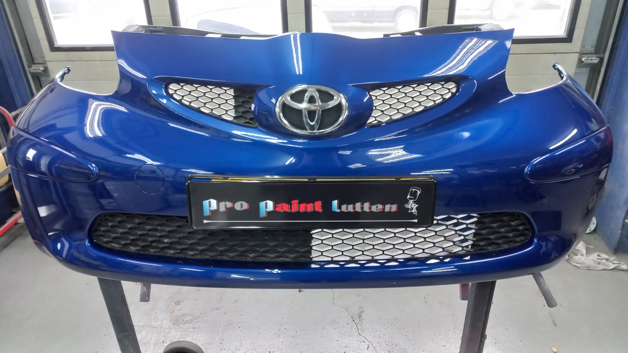 Een diepblauw gespoten voorbumper van een Toyota op een standaard, met een kentekenplaathouder van Pro Paint Lutten erin